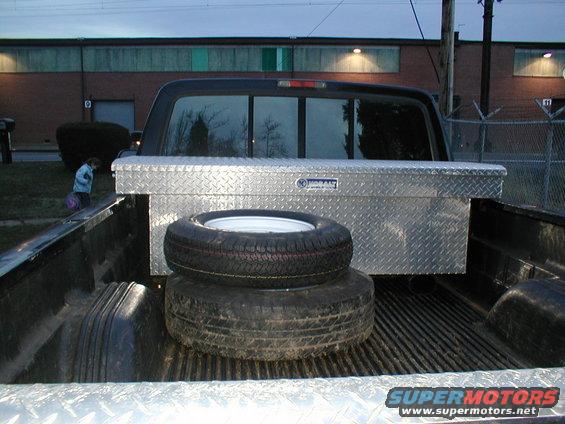 Pickup truck bed with diamond plate toolbox and two spare tires stacked. A person is visible in the background near a building