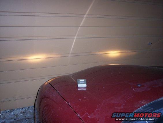 Close-up of a red car's hood with a silver hood ornament. Headlights are on, illuminating a garage door