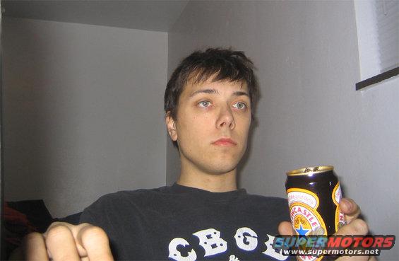 Man in a black t-shirt holding a Newcastle Brown Ale can, with a watermark for Supermotors.net