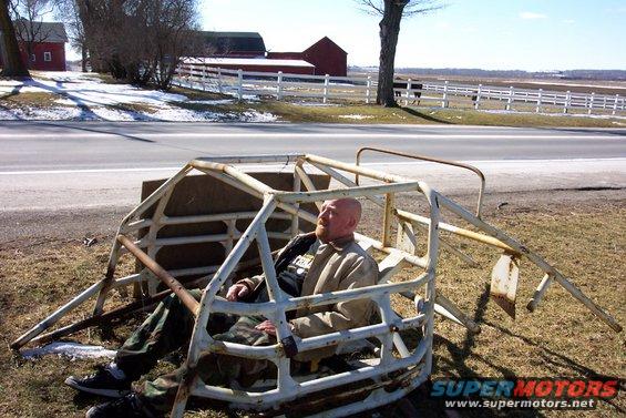 White metal roll cage with a man sitting inside, on grassy ground near a road and farm buildings