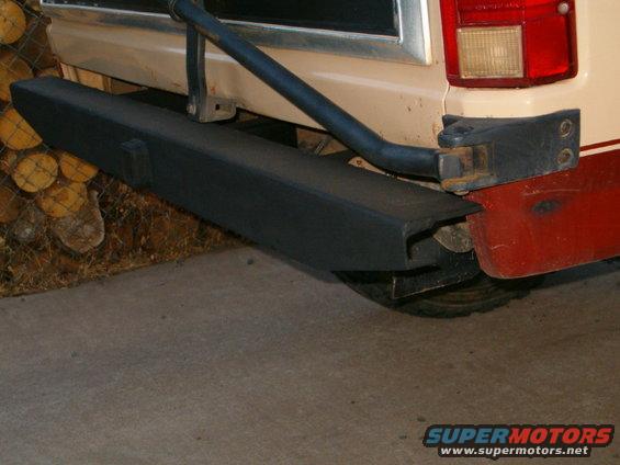 Rear bumper and trailer hitch of a tan and red vehicle, with logs in the background