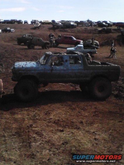 Muddy blue SUV with large tires parked in a dirt field with other vehicles in the background
