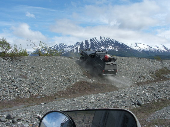SUV driving up a gravel hill with snow-capped mountains behind it. Shot from another vehicle's perspective