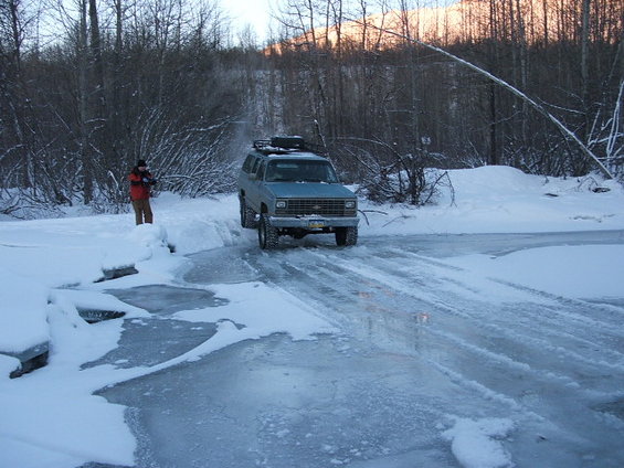 Light blue SUV with roof rack driving on icy road in snowy woods