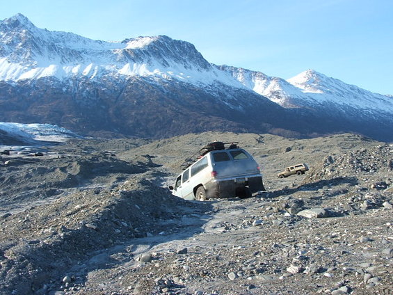 Silver SUV with roof rack stuck in rocky terrain with snow-capped mountains in background