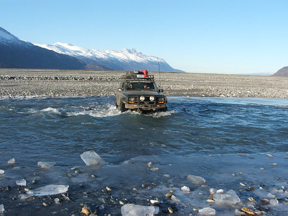 Dark SUV crossing icy river, front view. Snow-capped mountains behind
