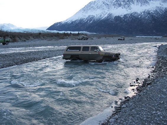 Two-tone Chevrolet Suburban with off-road tires driving through a river, mountains and glacier in background. Another vehicle is visible further back