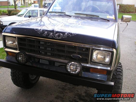 1986 Ford Bronco II front grille, headlights, and bumper with off-road lights. Another white vehicle is parked behind it