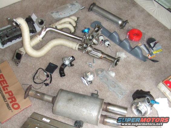Car parts including a turbocharger, exhaust manifold, and muffler are spread out on a carpeted floor