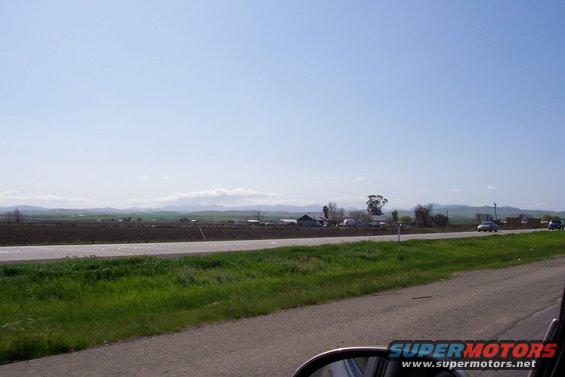 Two cars drive on a highway with a rural landscape and mountains in the background