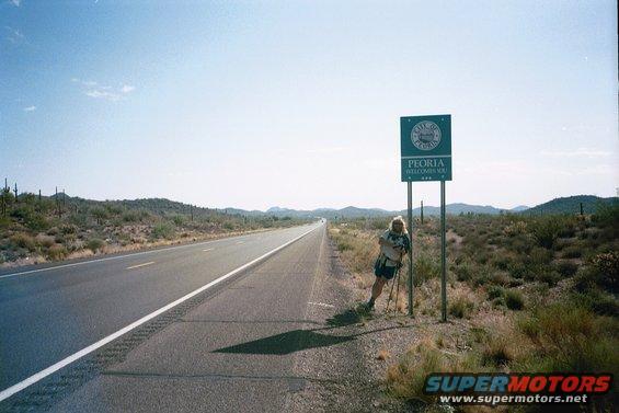 Person with hiking poles leaning on a sign that says 'City of Peoria Welcomes You' next to a highway in a desert landscape