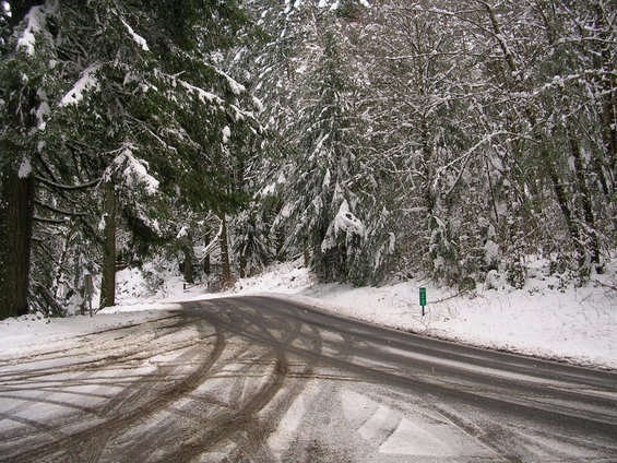 Winding asphalt road covered in patches of snow and tire tracks, surrounded by snow-dusted evergreen trees