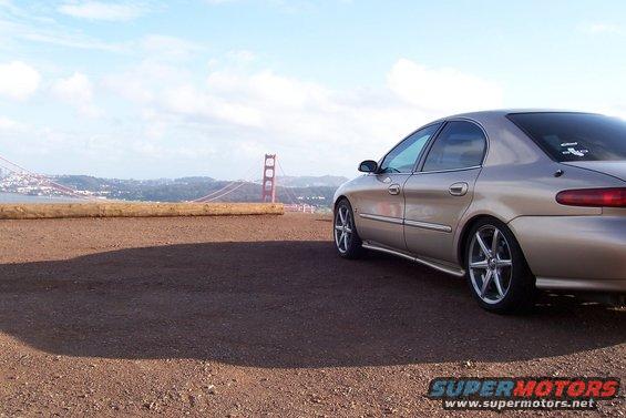 Tan sedan with aftermarket wheels parked on a dirt overlook, Golden Gate Bridge in background