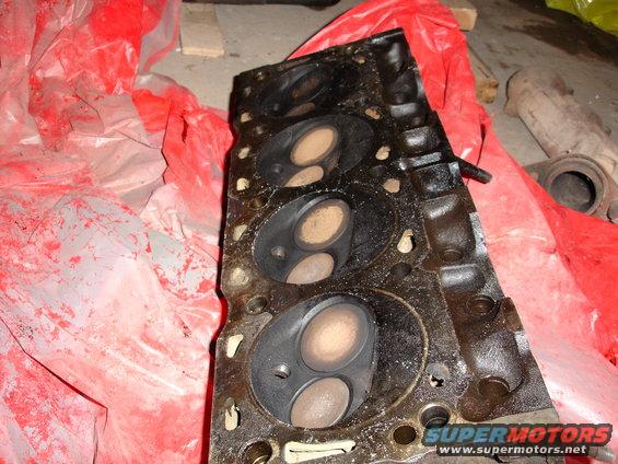 Black engine cylinder head with visible valves and combustion chambers, partially covered by red plastic sheeting
