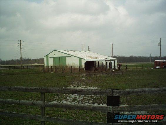 Barn with green doors and a red trailer in a muddy field under a cloudy sky
