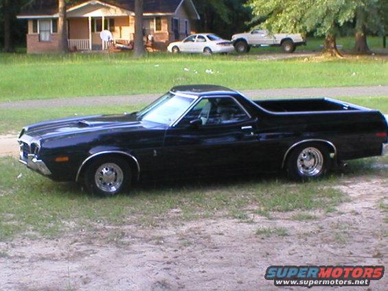 black pickup truck with a car cab, parked on grass in front of a house. Another truck and car are in the background