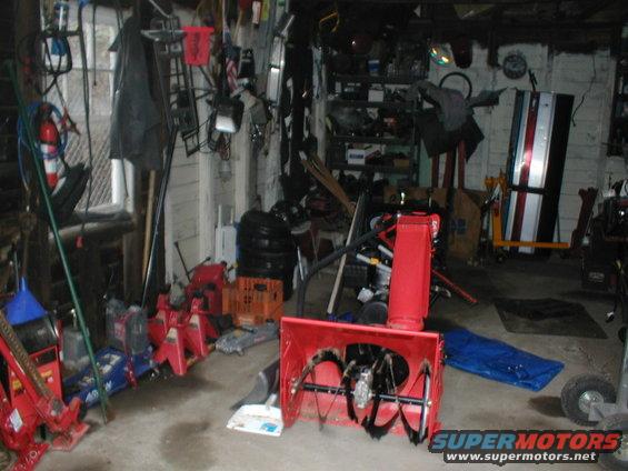 Cluttered garage interior with a red snowblower in the foreground, jack stands, tires, and various tools on shelves