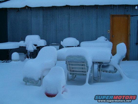 Patio furniture and tables covered in snow in front of a corrugated metal building with a yellow door