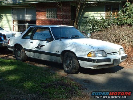 White 1987 Ford Mustang police car, 3/4 front view, parked in front of a house, showing "5.0" badge