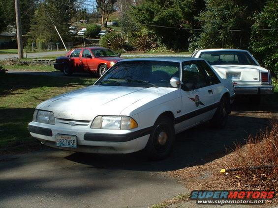 White 1987 Ford Mustang coupe, 3/4 front view, with a police-like decal on the door
