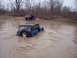 1974 Ford Bronco - Two off-road vehicles navigate a muddy terrain, with one vehicle partially submerged in water.
