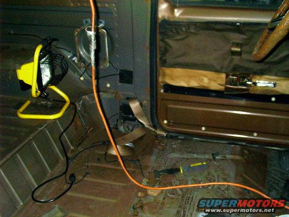 Interior of a vehicle with stripped floor, door panel, and steering wheel visible. A work light and orange extension cord are present