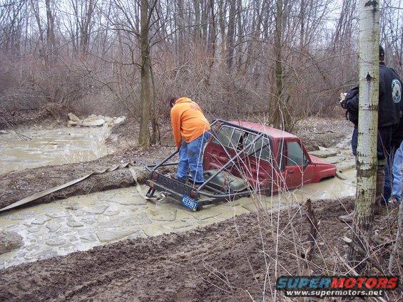 Red SUV stuck in a muddy ditch with a roll cage, people trying to pull it out