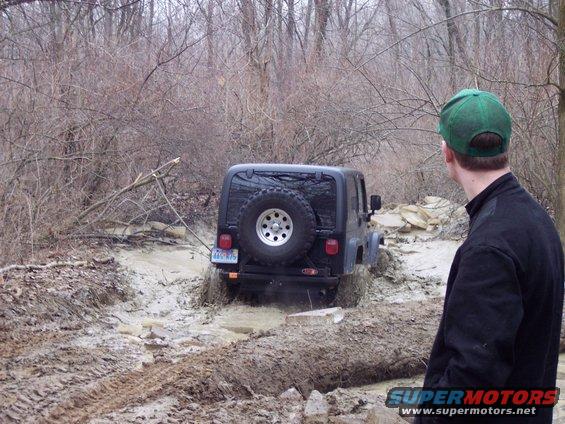 Jeep Wrangler driving through a muddy trail in the woods. A person in a green hat watches