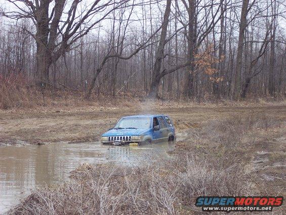 Blue Jeep Grand Cherokee stuck in a muddy puddle with trees in the background