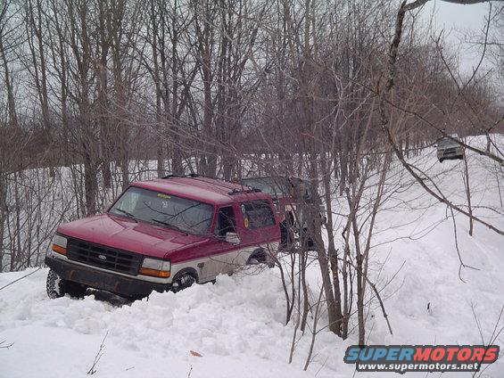 Red and tan Ford Bronco stuck in deep snow, another vehicle visible on a snowy hill in the background