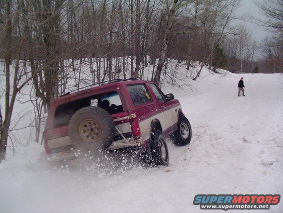 Red and white Ford Bronco driving in snow, kicking up powder. Spare tire mounted on back