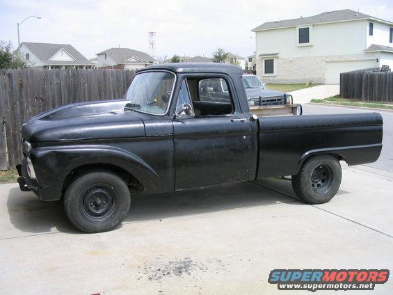 Side view of a black 1965 Ford F-100 pickup truck parked on a driveway. Another truck is visible in the background