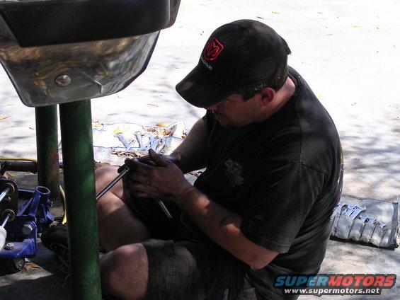 Man in black t-shirt and cap works on a metal vehicle part with a socket wrench