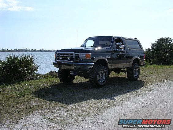 Black lifted Ford Bronco parked on a dirt road near water, with tan side trim and chrome wheels
