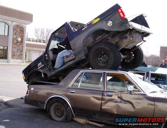 Black Ford Bronco on top of a damaged brown sedan, driver visible in Bronco