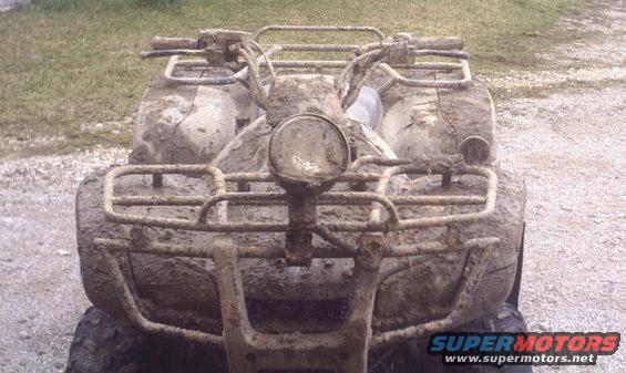 Mud-covered quad bike, front view. Headlight and handlebars are visible