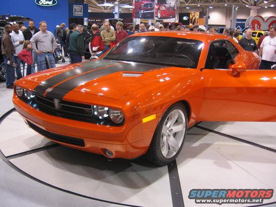Orange Dodge Challenger with black racing stripes, driver door open, on display at an auto show