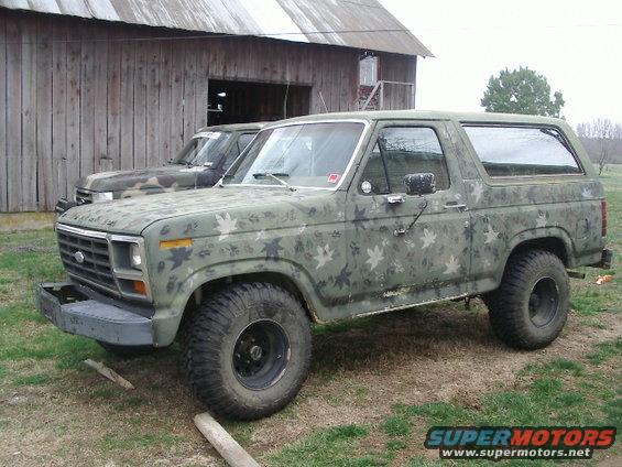 1984 Ford Bronco with camouflage paint parked in front of a barn, another vehicle behind it