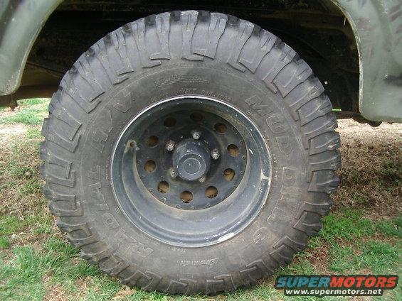 Off-road tire on a black rim, showing part of a green vehicle body and suspension. Text on tire reads "MUD-TERRAIN T/A"