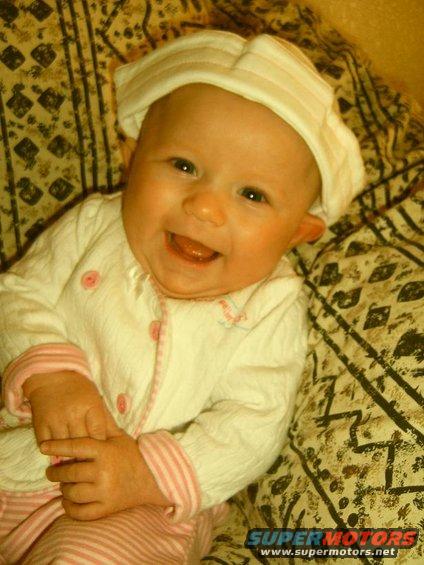 Smiling baby wearing a white hat and outfit, lying on a patterned blanket