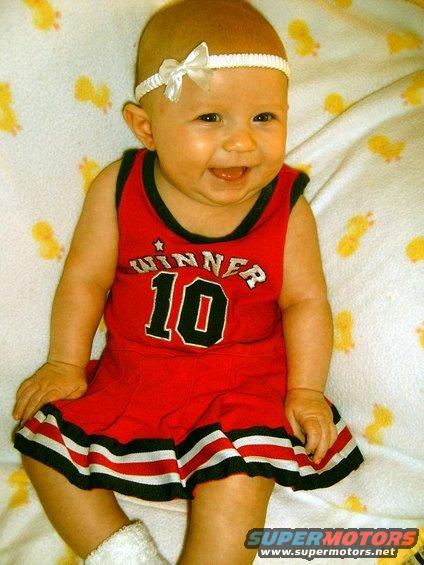 Baby in red 'Winner 10' outfit with white headband and bow, lying on a yellow duck-patterned blanket