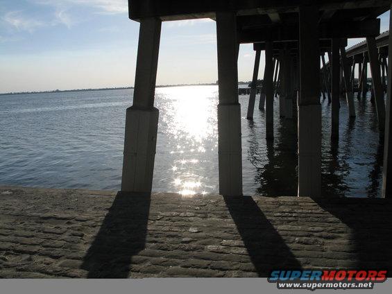 Concrete pier pilings rise from the water under a bright sun, casting long shadows on a stone walkway