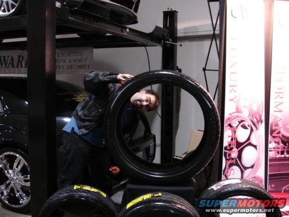 Guy peeking through a tire at a car show, with other tires and displays around