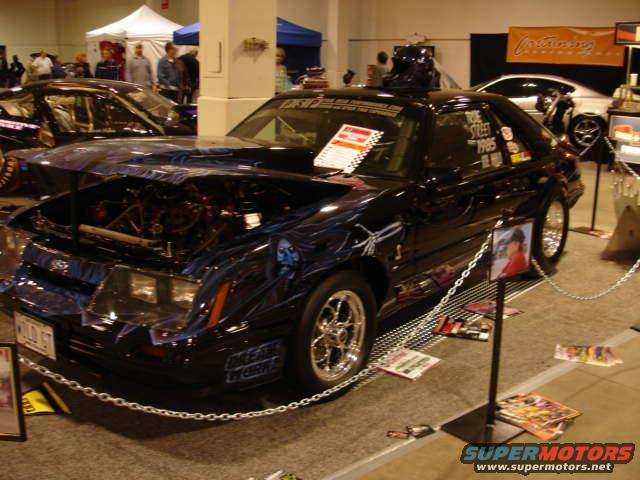 Black customized Ford Mustang with airbrushed graphics, hood open showing engine, custom wheels, and a "1985" badge on the side