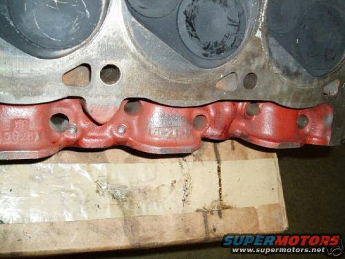 Close-up of an engine cylinder head with an intake manifold attached. Part numbers are visible on the manifold