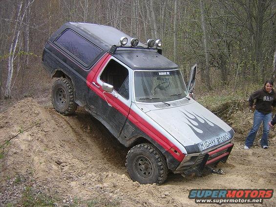 Ford Bronco with flame decals and "MIGHTY BRONCO" on the hood, driving up a dirt hill. A woman watches