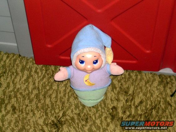 Toy doll with blue hat and purple shirt with yellow moon, on green carpet in front of red structure