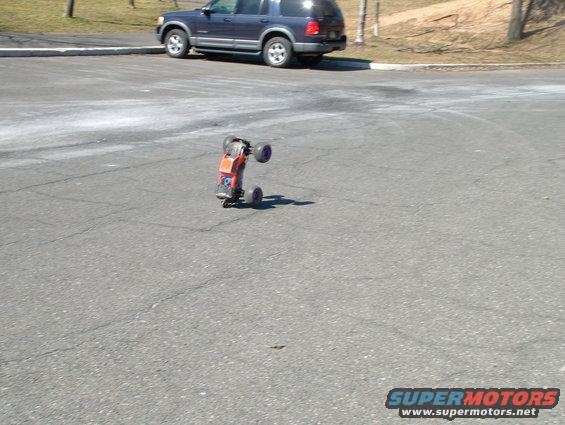 Orange remote control car tipped on its side on asphalt, with a blue SUV in the background