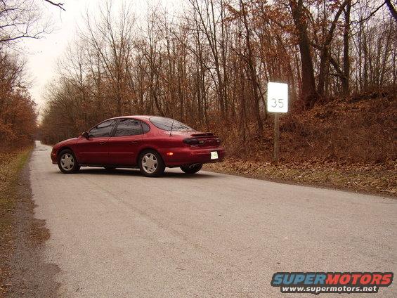 Red 1997 Ford Taurus sedan driving on a paved road next to a speed limit sign