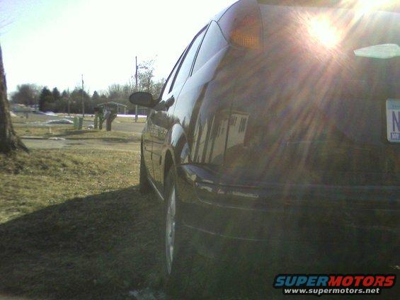 Black hatchback car from rear-side angle, parked on grassy area. Sun glare obscures some details